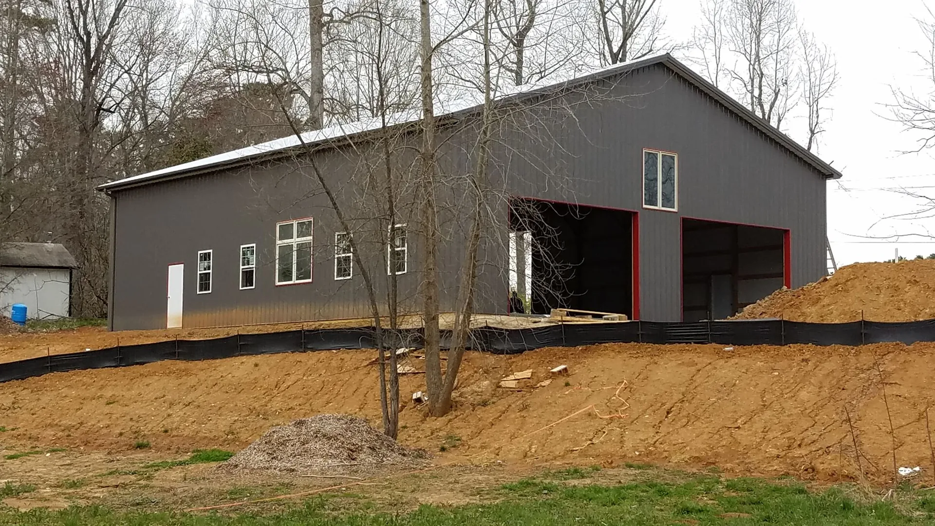 A new gray industrial building with a large open bay door, surrounded by a mound of bare earth, under a cloudy sky.
