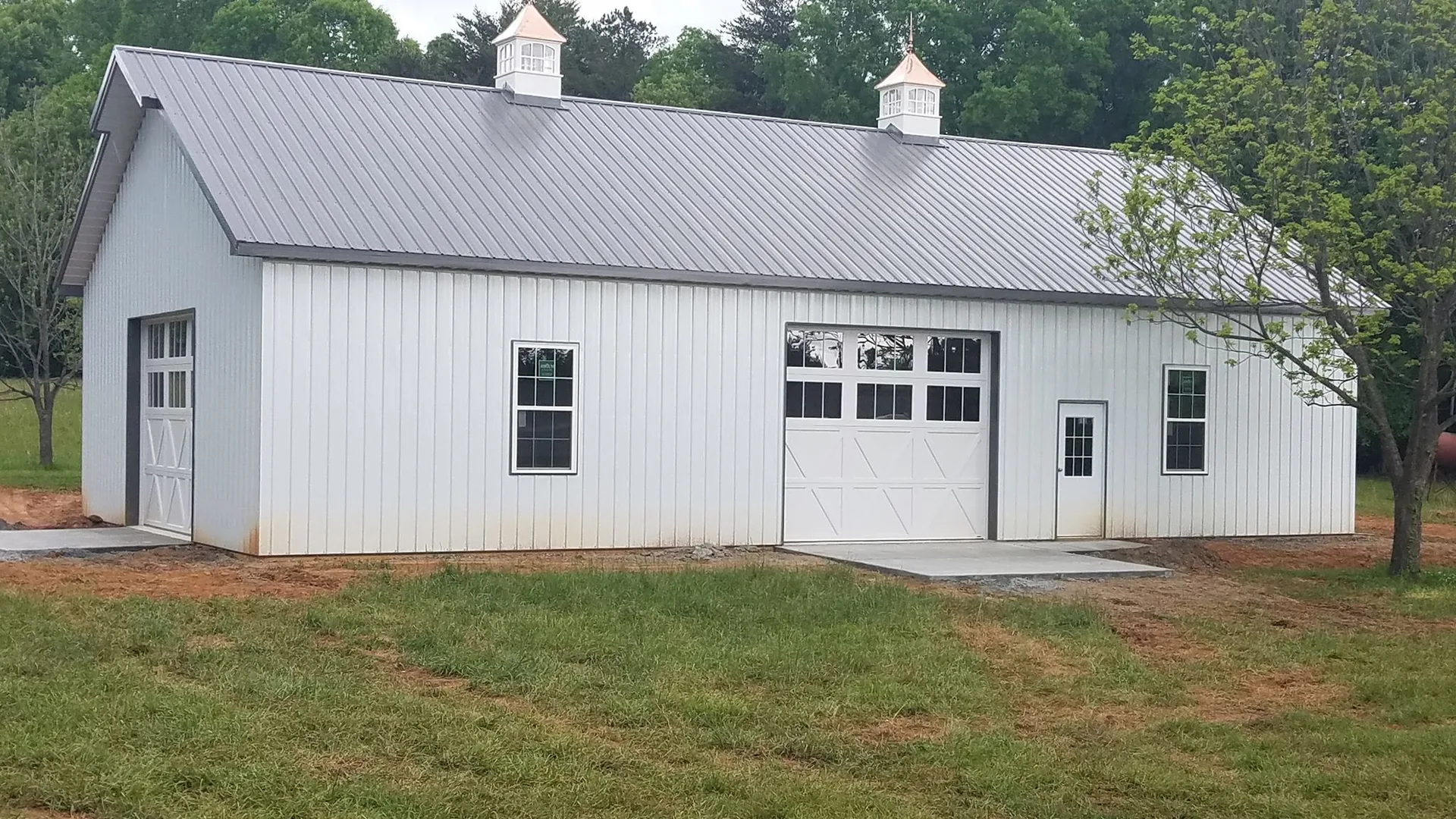 A white metal building with a gable roof, two cupolas, and large garage doors surrounded by a grassy area.
