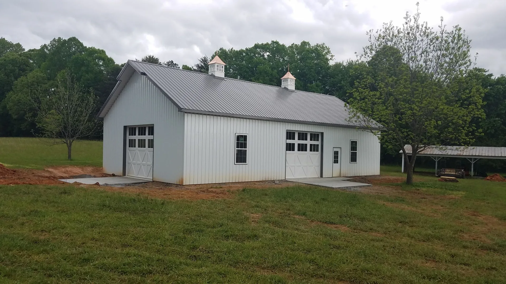 A large white barn with a metal roof and double doors sits on a grassy field with trees in the background.