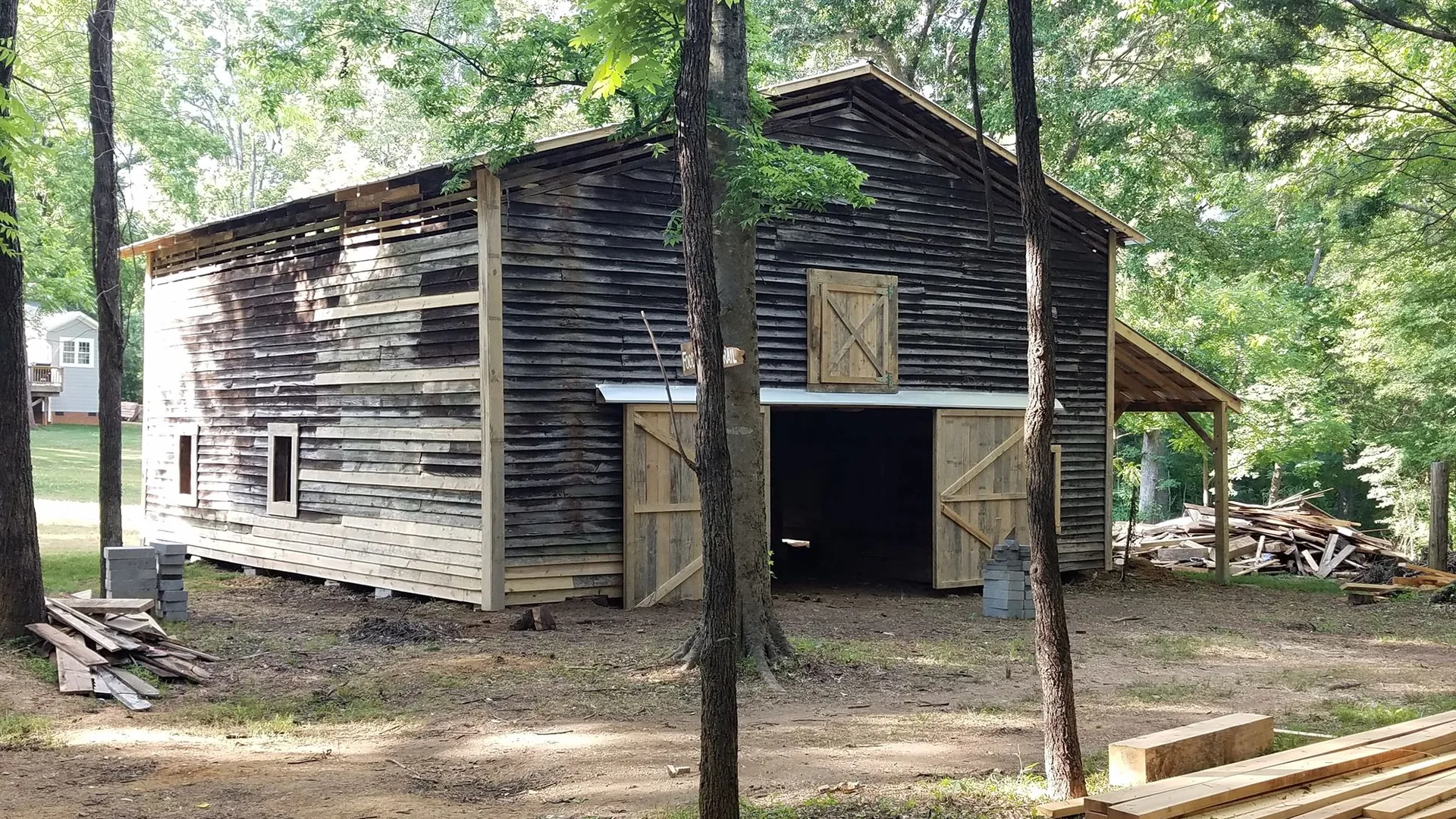 An old wooden barn with an attached shed stands surrounded by trees with lumber scattered in the vicinity.