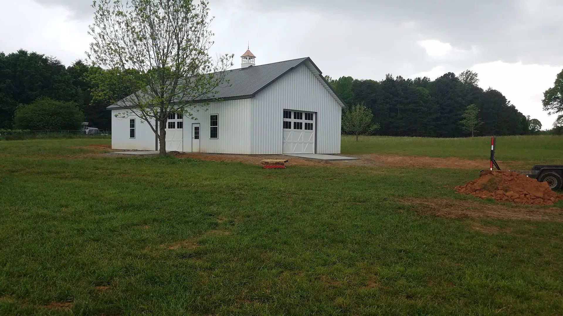 A white rural building with a large door and a smaller adjacent door, surrounded by greenery under a cloudy sky.