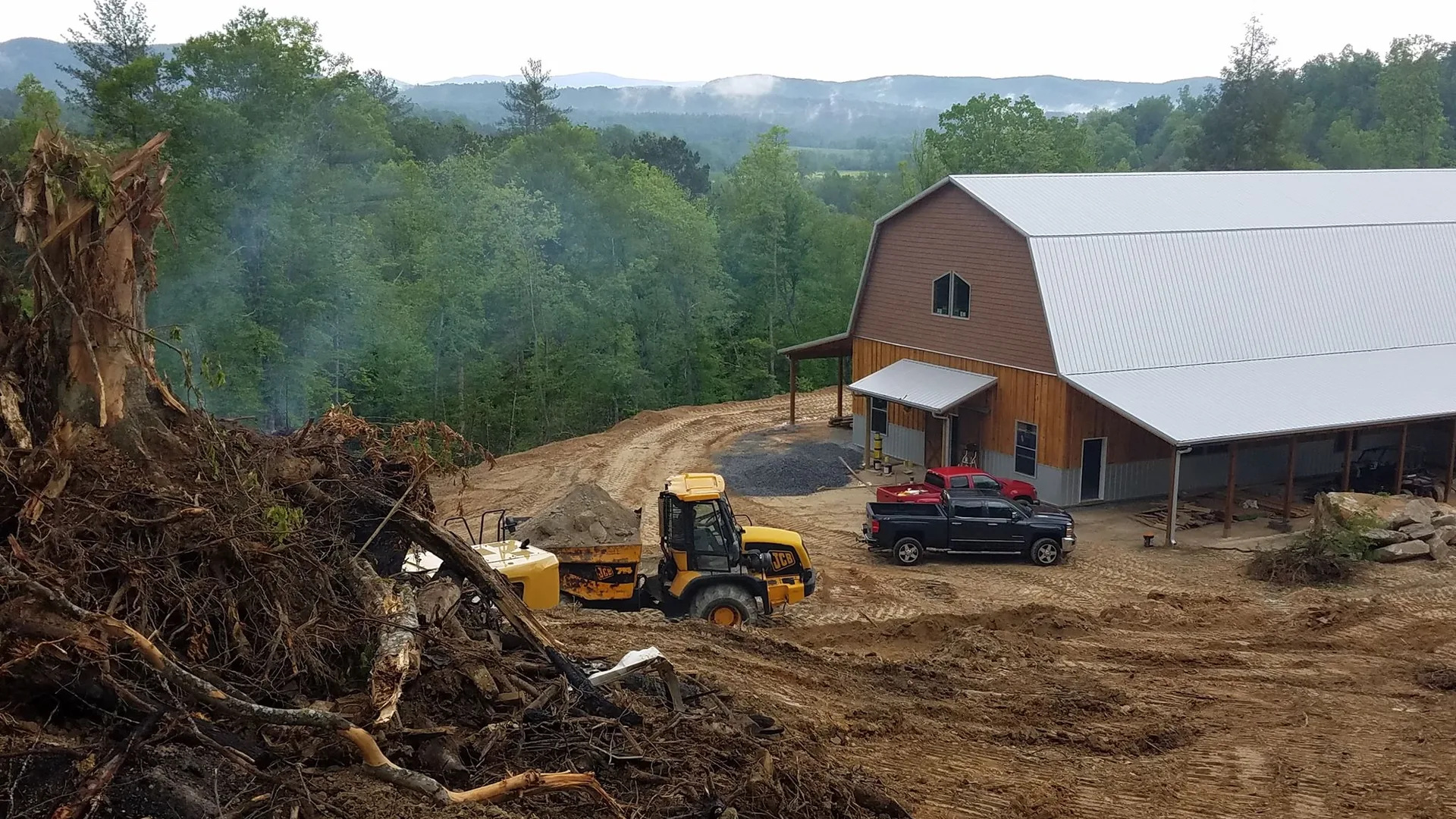 A construction site with heavy machinery, a pickup truck, and a large building against a backdrop of forested hills.