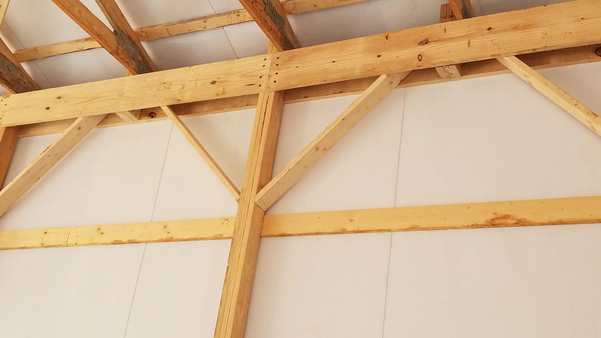 The image shows a close-up view of a wooden roof structure under construction with visible beams and joints against an insulated wall.