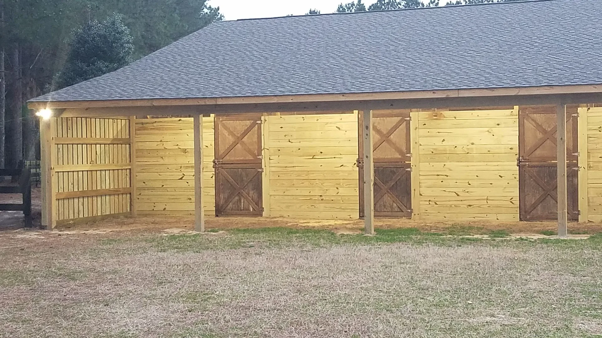 A wooden stable with multiple stalls and a lit corner, surrounded by a grassy area and trees in the background.