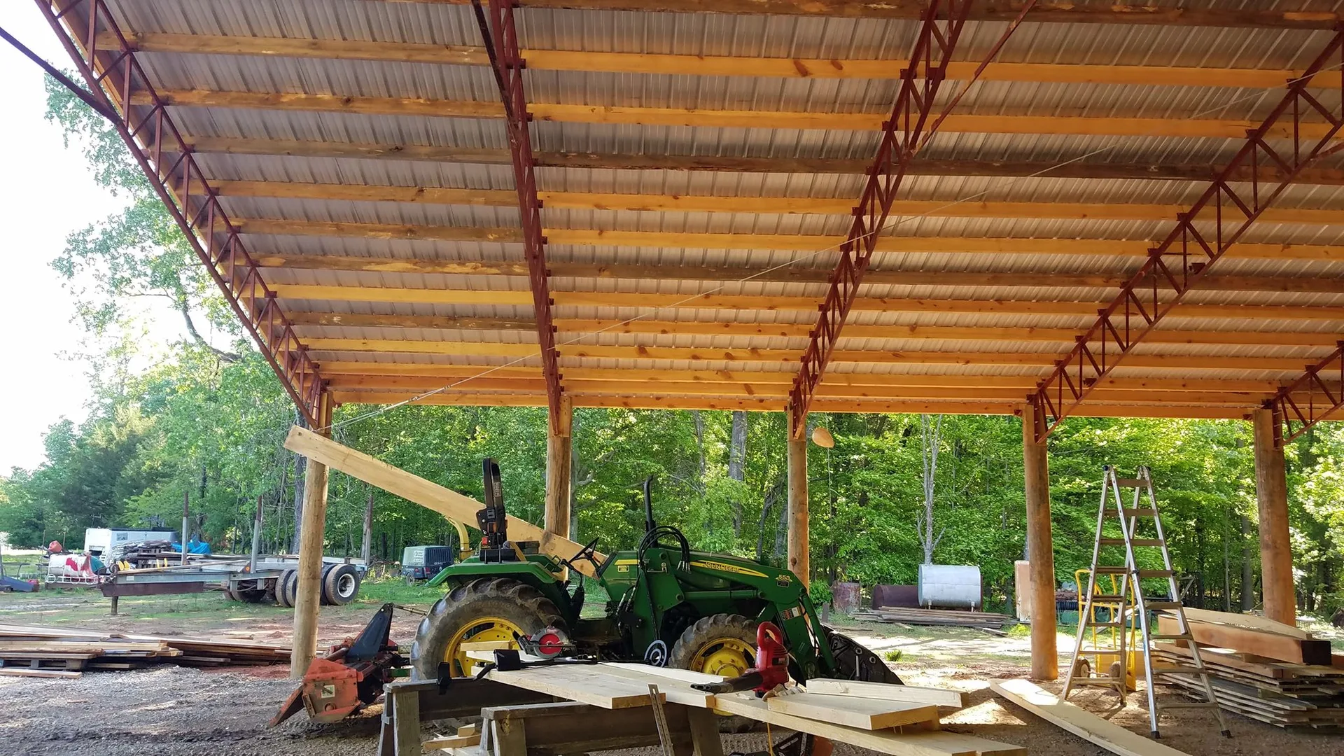 A tractor under a partially constructed metal shelter surrounded by building materials and trees in the background.