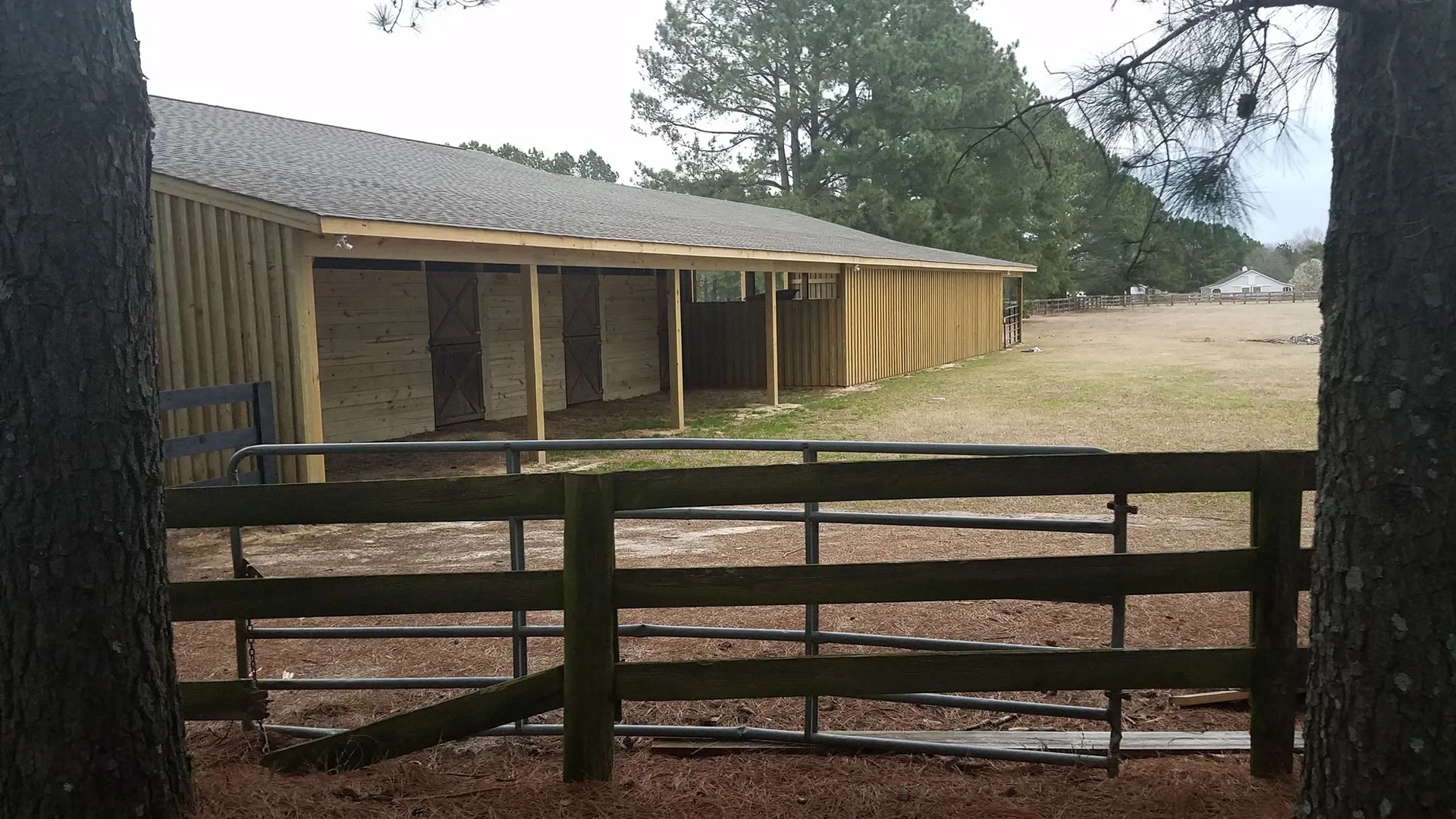 A wooden stable with a fenced paddock surrounded by trees on a cloudy day.