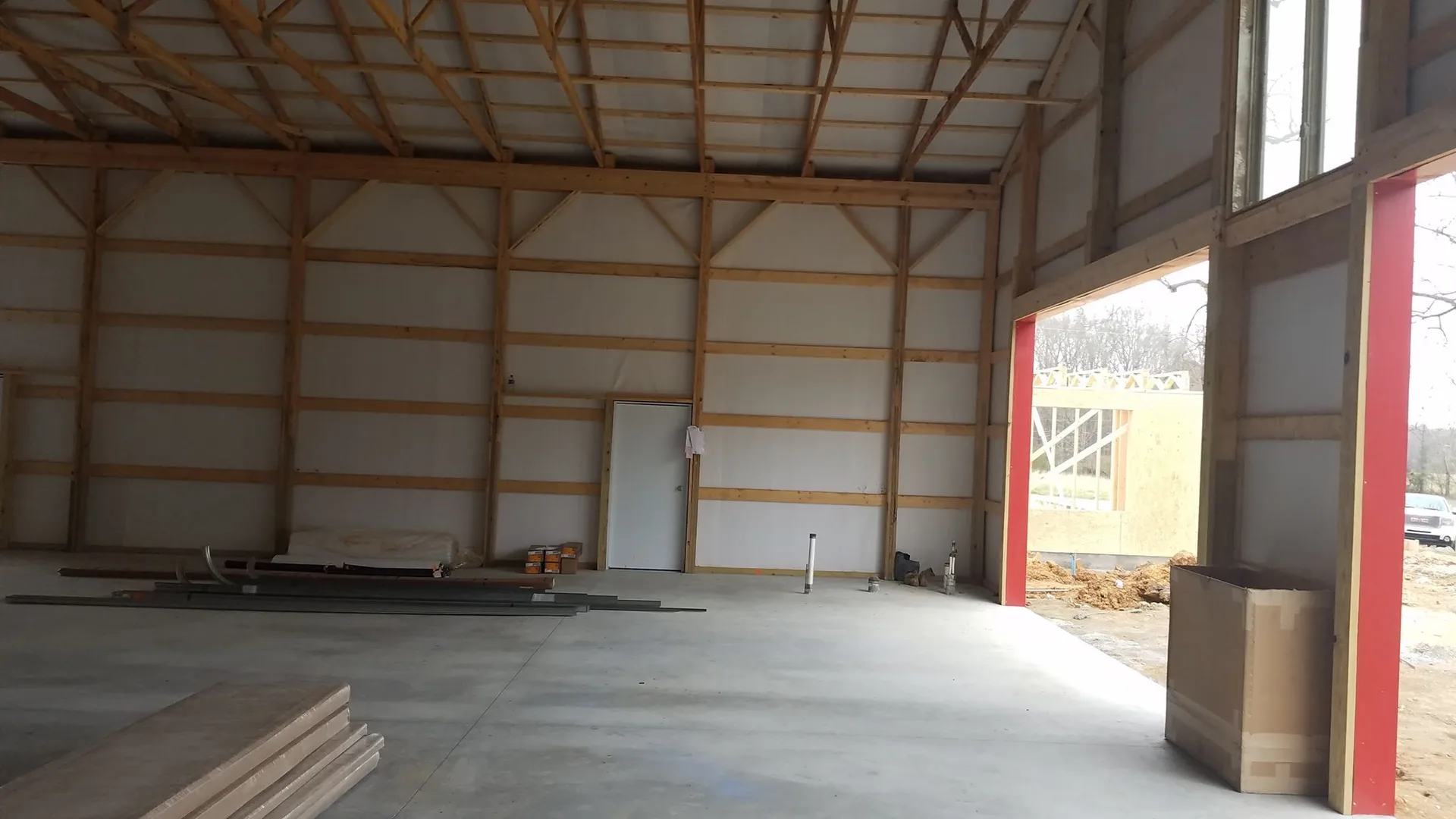 An empty industrial building interior with wooden beams, a concrete floor, large garage doors, and construction materials scattered around.