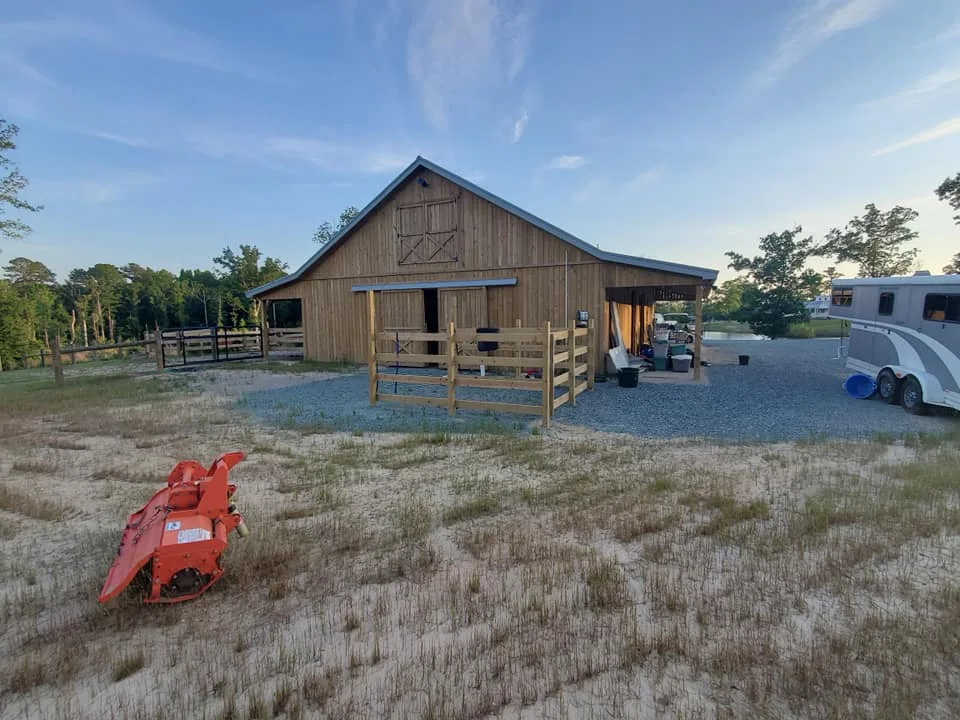 A rustic barn stands amid a tranquil field with an orange tractor attachment in the foreground and a horse trailer parked to the side.
