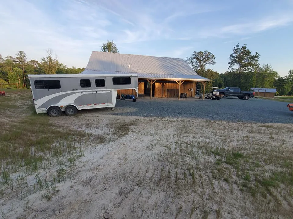 A white horse trailer is parked outside a large barn with a metallic roof, with a pickup truck and an ATV nearby on a gravel area.