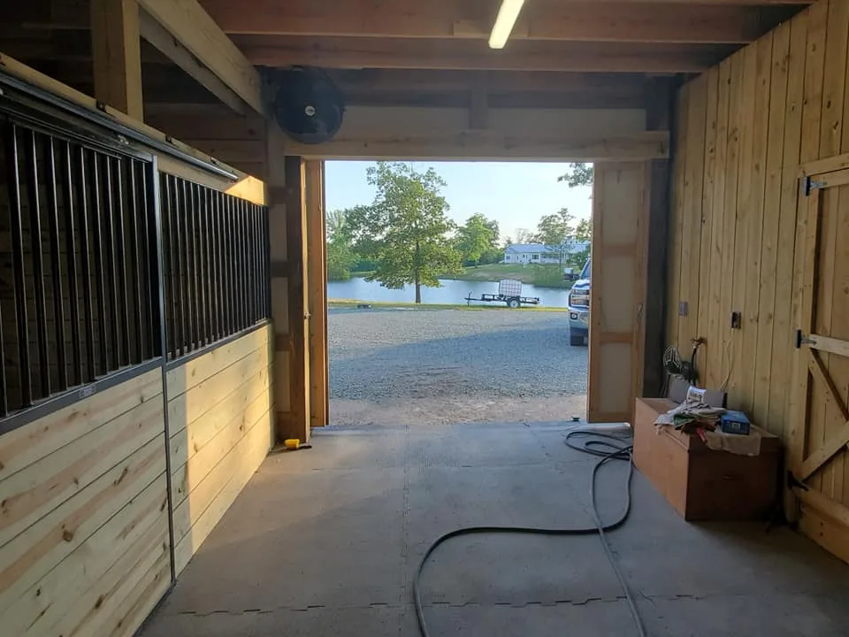 A spacious wooden barn interior with a view of an outdoor area featuring a water body and a parked vehicle.