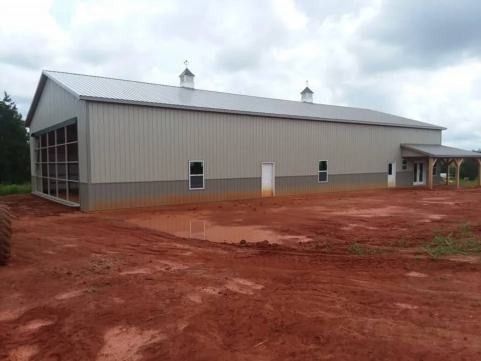 A large metal barn with a sloped roof and multiple vents sits on a plot of reddish soil under a cloudy sky.