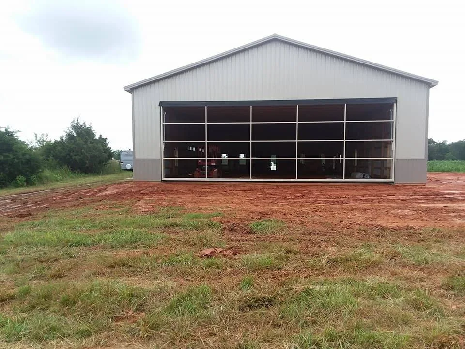 A large industrial metal building with an open grid-style door and a dirt ground in the foreground under a cloudy sky.