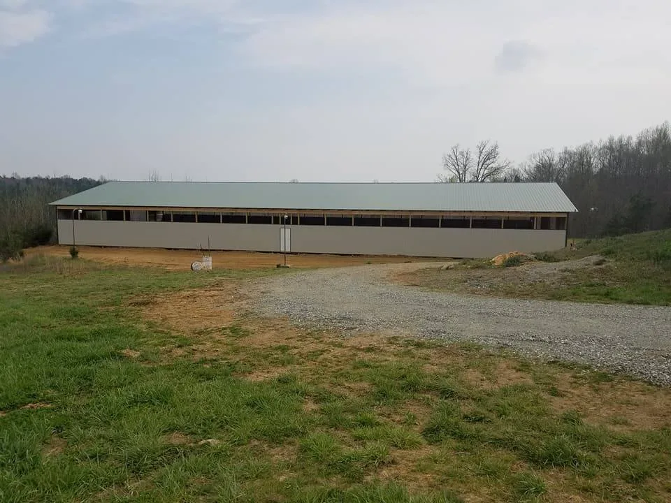 A long, single-story building with a brown metal roof and numerous windows, situated in a grassy field under a cloudy sky.