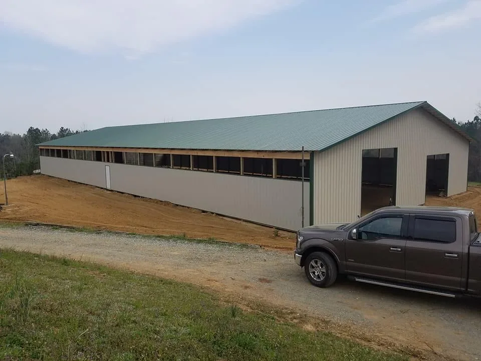 A large metal agricultural building with a green roof and a pickup truck parked beside it on a cleared dirt area.