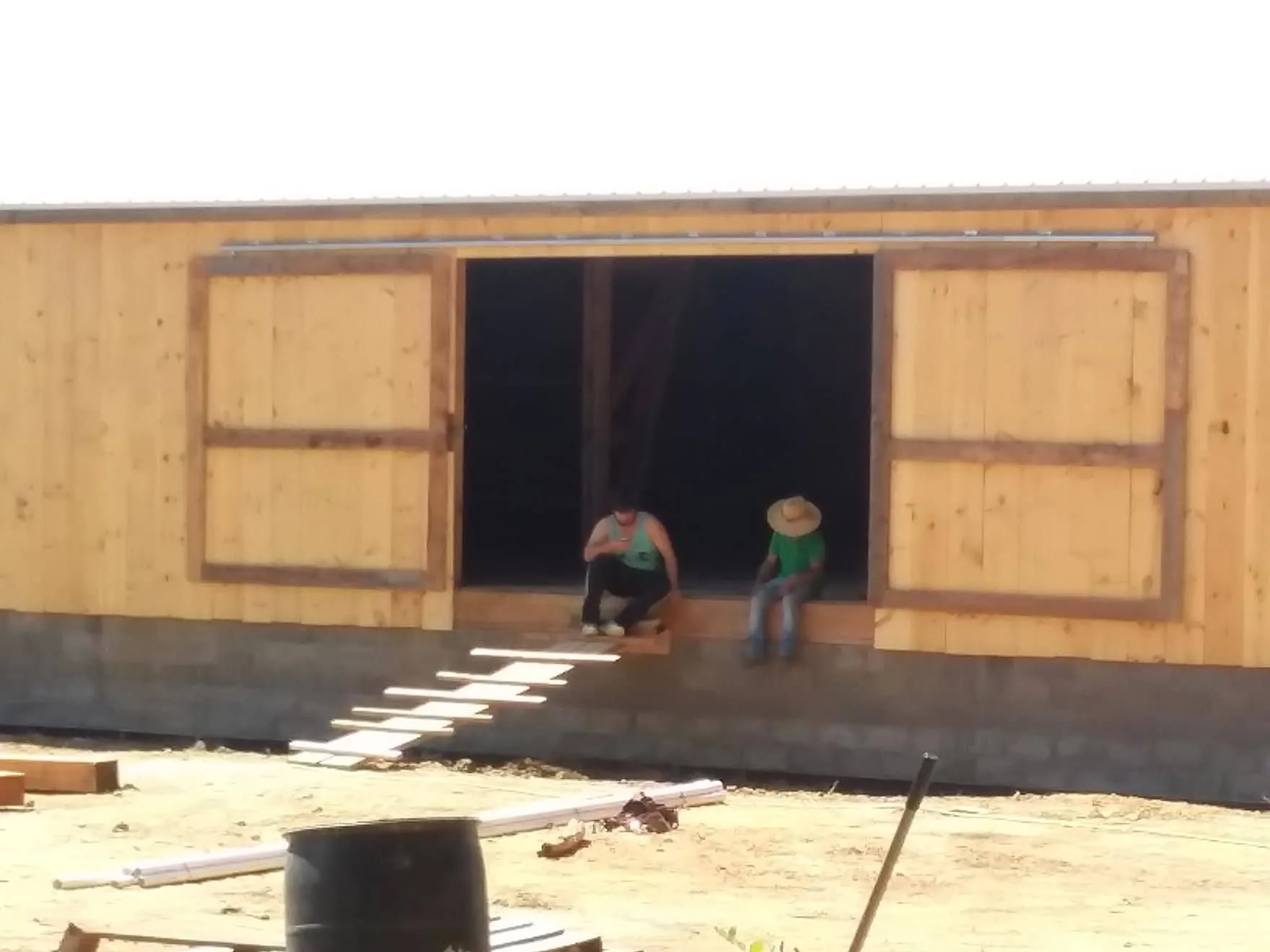 Two people are sitting at the entrance of a wooden structure under construction on a sunny day.