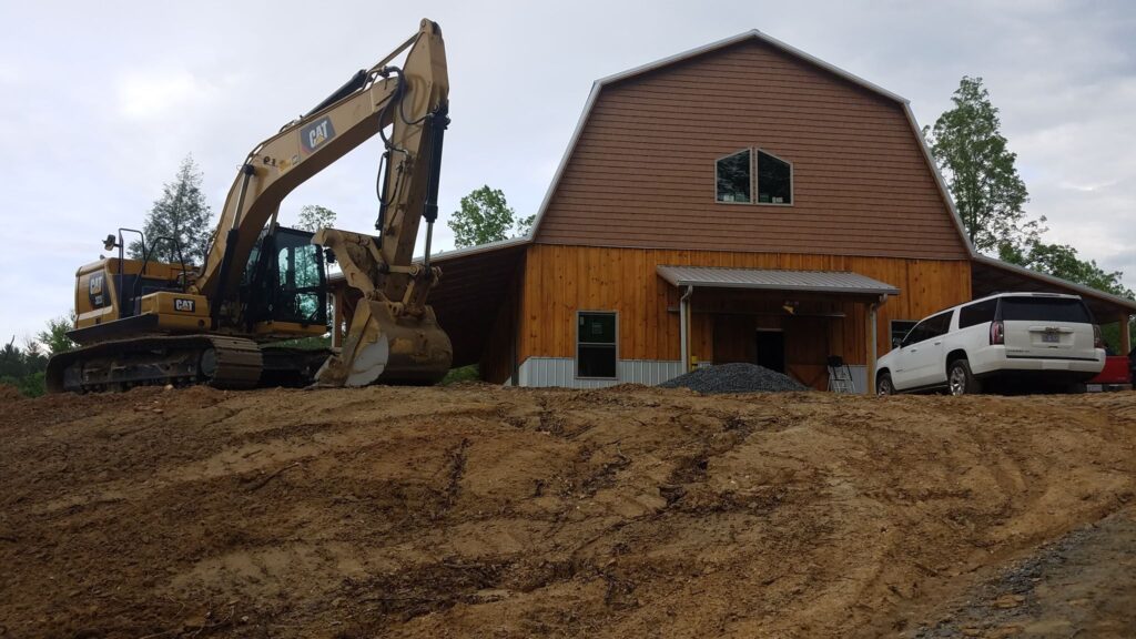 A construction scene with an excavator parked next to a new barn house and an SUV on a dirt terrain.