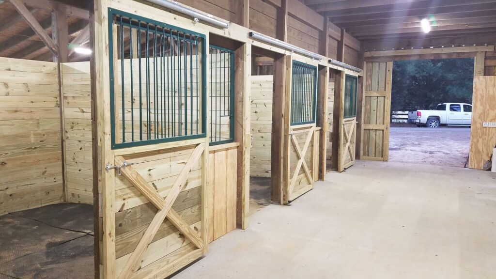 A row of empty wooden horse stalls in a stable with open doors and a vehicle parked outside.
