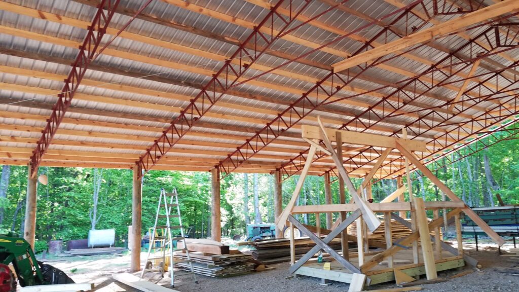 The image shows a construction site with a metal roof structure, wooden supports, a ladder, and various building materials scattered around.
