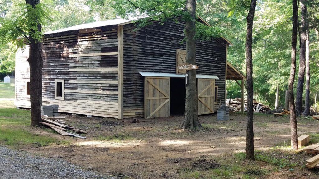 A rustic wooden blacksmith shop surrounded by trees with lumber scattered around it.