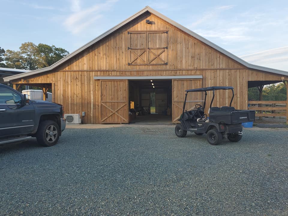 A wooden barn with open doors, flanked by a pickup truck and a utility vehicle, with the sky transitioning to dusk in the background.