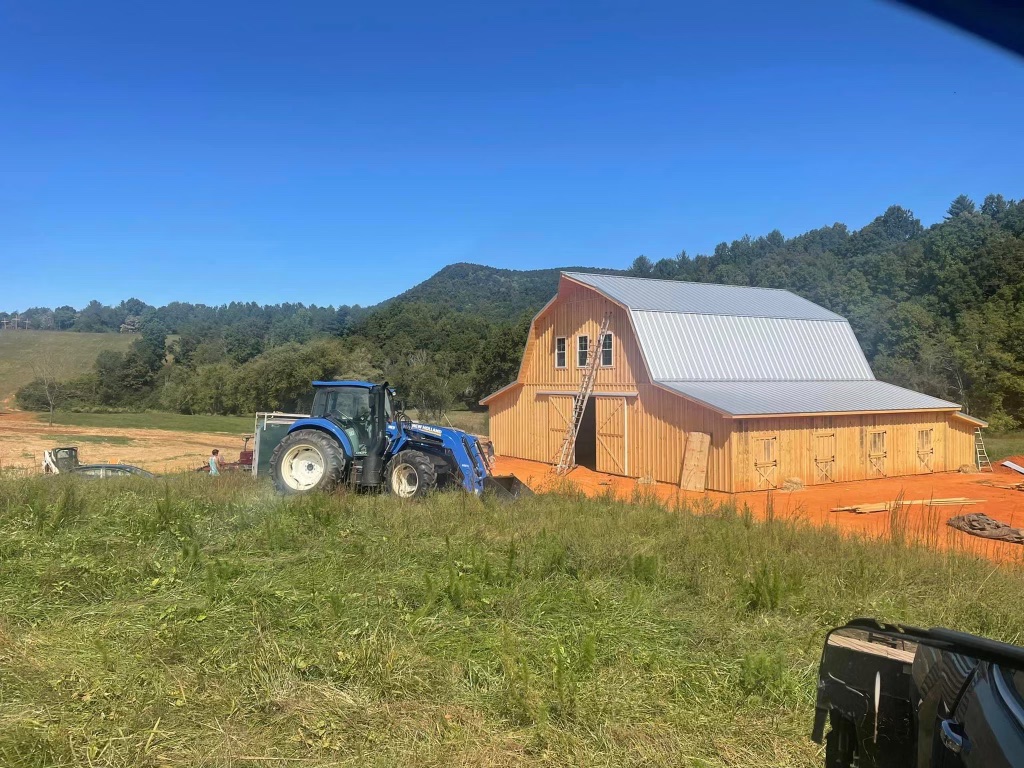 A blue tractor in front of a newly constructed wooden barn surrounded by a grassy field with hills in the background.