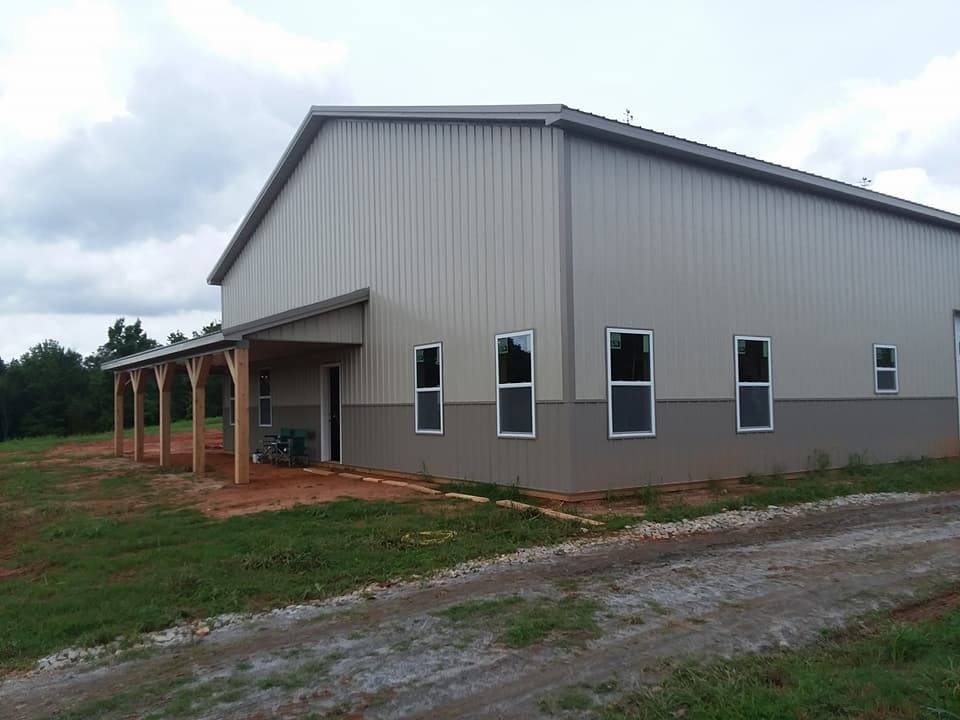 A new gray metal building with a covered porch under construction in a dirt-covered area with cloudy skies.