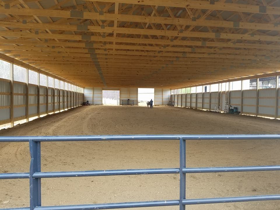 An indoor horse riding arena with a sandy floor, wooden ceiling, and a person in the distance.